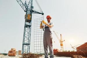 Crane is at background. Young construction worker in uniform is busy at the unfinished building photo