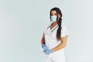 Standing against white background. Young female doctor in uniform is indoors photo