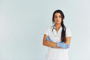 Standing against white background. Young female doctor in uniform is indoors photo