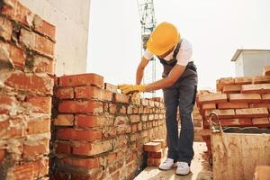 Using bricks. Young construction worker in uniform is busy at the unfinished building photo