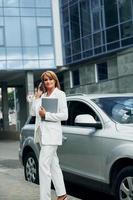 With phone in hand. Woman in formal wear standing outdoors in the city at daytime photo