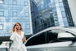 Having conversation by the phone. Woman in formal wear standing outdoors in the city at daytime photo