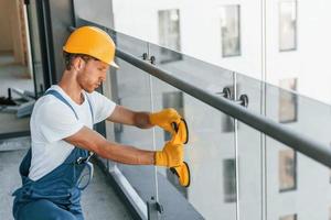 Using tool on glass. Young man working in uniform at construction at daytime photo