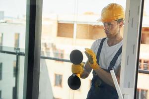 Using tool on glass. Young man working in uniform at construction at daytime photo