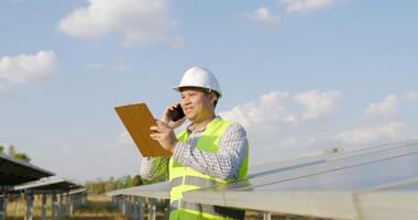 Slow motion shot, Young engineer in white helmet holding checking board in hand standing and talking on smartphone while working at solar farm in background video