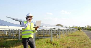 Slow motion shot, Young engineer in white helmet holding laptop computer in arm standing and talking on smartphone while working at solar farm in background video