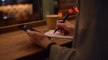 kvinna använda sig av smartphone i de Kafé och skriva anteckningar i anteckningsbok, långsam rörelse video