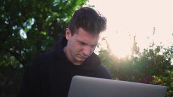 chico trabaja en una computadora portátil en un parque de la ciudad con el telón de fondo de la vegetación. hombre independiente usa cuaderno para trabajo al aire libre. concepto de trabajo remoto. camara lenta. video