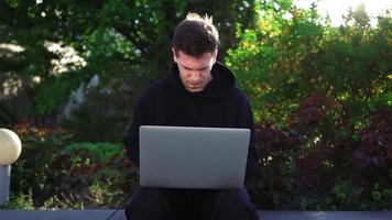 guy travaille sur un ordinateur portable dans un parc de la ville sur fond de verdure. un pigiste masculin utilise un ordinateur portable pour le travail en extérieur. notion de travail à distance. ralenti. video