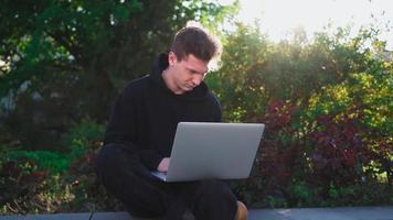 chico trabaja en una computadora portátil en un parque de la ciudad con el telón de fondo de la vegetación. hombre independiente usa cuaderno para trabajo al aire libre. concepto de trabajo remoto. camara lenta. video