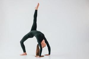 Against white background. Young woman in sportive clothes doing gymnastics indoors photo