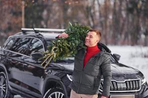 sosteniendo abeto verde en las manos. el hombre está al aire libre cerca de su auto. concepción de las vacaciones foto