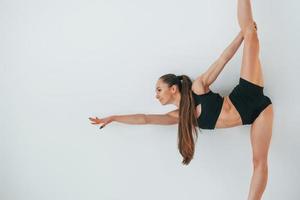 Standing against white background. Young woman in sportive clothes doing gymnastics indoors photo