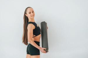 Standing against white background. Young woman in sportive clothes doing gymnastics indoors photo