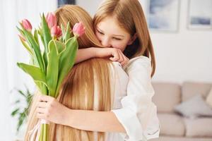 Embracing each other. Young mother with her daughter is at home at daytime photo