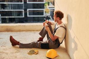 apoyado en la pared. joven que trabaja en uniforme en la construcción durante el día foto