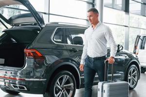 Conception of tourism. Man in formal clothes is with luggage indoors near the car photo