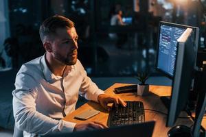 Working at evening time. Young businessman in formal clothes is in office with multiple screens photo