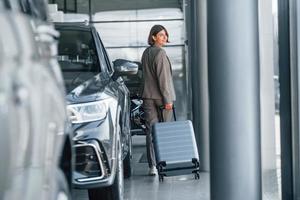 Holding luggage. Woman is indoors near brand new automobile indoors photo
