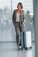 Against wall with reflection. Woman with luggage is standing indoors. Conception of tourism photo