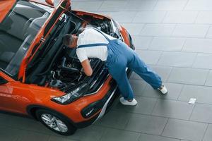 In blue uniform. Man is repairing broken automobile indoors photo