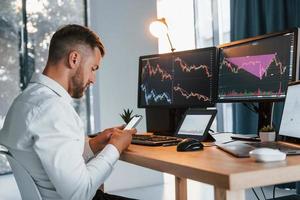 Young businessman in formal clothes is in office with multiple screens. Conception of exchange photo