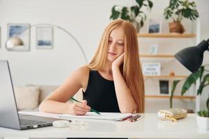 Sitting by the table. Female teenager with blonde hair is at home at daytime photo