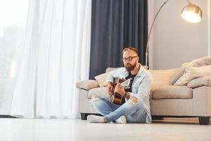 Sitting near the sofa. Man in casual clothes and with acoustic guitar is indoors photo