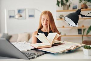 con libro y laptop. una adolescente con cabello rubio está en casa durante el día foto