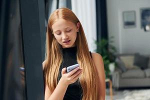 Standing with phone. Female teenager with blonde hair is at home at daytime photo
