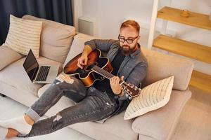 Having good time practicing. Man in casual clothes and with acoustic guitar is indoors photo