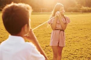 Boy with girl having fun by using tin can telephone while standing outdoors on the field photo