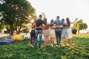 vista desde atrás. grupo de jóvenes tienen una fiesta en el parque durante el día de verano foto