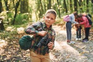 Boy with binoculars standing in front of his friends. Kids in green forest at summer daytime together photo
