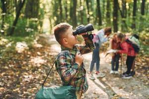 Boy with binoculars standing in front of his friends. Kids in green forest at summer daytime together photo