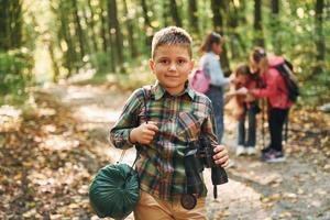 chico con binoculares parado frente a sus amigos. niños en el bosque verde durante el día de verano juntos foto