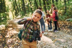 chico con binoculares parado frente a sus amigos. niños en el bosque verde durante el día de verano juntos foto