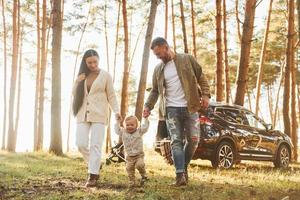 Against black automobile. Happy family of father, mother and little daughter is in the forest photo