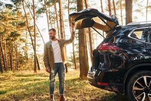 Near the car. Man in jeans is outdoors in the forest with his black colored automobile photo