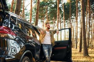 With his car. Man in jeans is outdoors in the forest with his black colored automobile photo