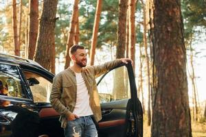 con su coche el hombre en jeans está afuera en el bosque con su automóvil de color negro foto