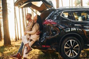 With black colored automobile. Happy family of father, mother and little daughter is in the forest photo