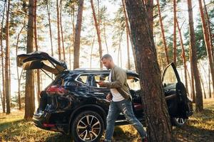 Near the car. Man in jeans is outdoors in the forest with his black colored automobile photo