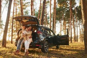Leaning on the car. Happy family of father, mother and little daughter is in the forest photo