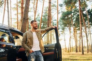 With his car. Man in jeans is outdoors in the forest with his black colored automobile photo