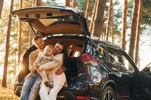 con automóvil de color negro. feliz familia de padre, madre e hija pequeña está en el bosque foto