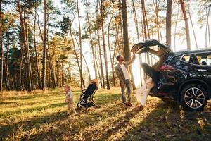 With black colored automobile. Happy family of father, mother and little daughter is in the forest photo