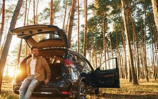 con su coche el hombre en jeans está afuera en el bosque con su automóvil de color negro foto