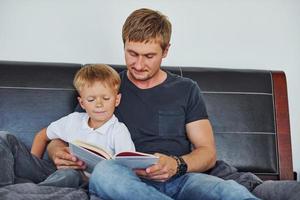 With book in hands. Father and son is indoors at home together photo