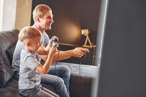 Holding video game controllers. Father and son is indoors at home together photo